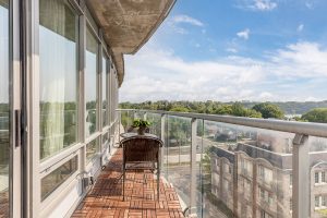 Balcony with wooden floor and glass railing overlooking cityscape under blue sky.